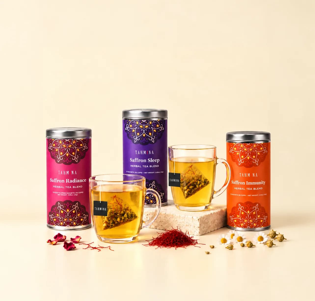 Saffron Radiance, Saffron Sleep and Saffron Immunity tins with two glass mugs of tea, saffron threads, rose petals and chamomile flowers on a cream ground